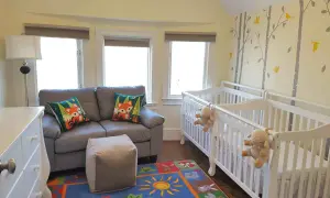 A charming nursery with two white cribs, perfect for nursery sets for twins. A grey sofa with fox-themed pillows and a colorful rug complete the cozy space.