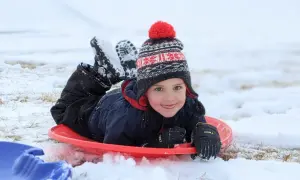 A happy child in a winter hat and snowsuit lies on a red sled, ready for fun. Perfect for things to do with baby in winter, like sledding!