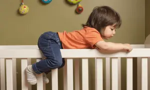 A toddler in jeans and an orange shirt is climbing over the side of a crib, with their legs dangling, as if preparing for dropping crib mattress to floor.