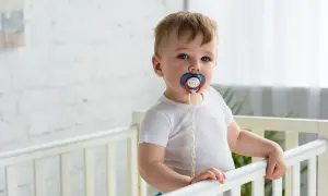 A baby in a crib with a pacifier. Some wonder if a pacifier can cause speech delay, but this little one seems content.