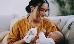 A mother smiles while paced feeding her newborn with a bottle. The baby looks up at her, reaching out a hand.