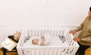 A happy baby rolls around in a white baby crib twin. A man smiles as he looks into the crib.
