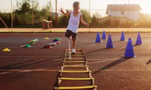 A young boy practices gross motor activities for 5 year olds, stepping through an agility ladder on a sports field with cones.