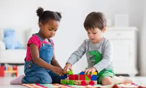 Two children engage in parallel play, building a colorful wooden train with blocks.