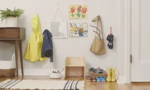 Montessori entryway ideas: a yellow raincoat, denim jacket, and artwork hang on the wall. A small wooden stool and baskets hold shoes and boots.