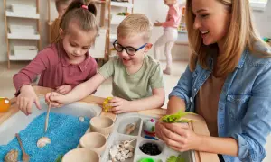 Children explore blue sand and shells in a sensory table, guided by a smiling teacher.