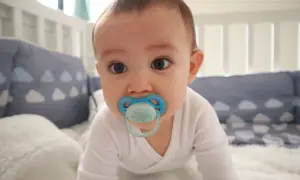 A baby with a blue orthodontic pacifier in a crib. Are orthodontic pacifiers better for developing smiles?