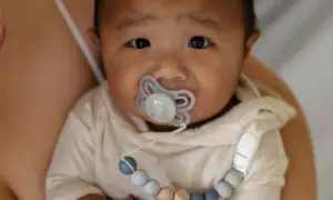 A baby with a pacifier, possibly experiencing pacifier nipple confusion, looks into the camera while being held by a parent.
