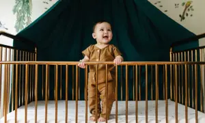 A happy baby stands in a golden crib, holding the bars. What do you need for a crib? A safe space, a soft mattress, and a smiling baby!
