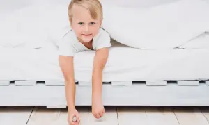 A young boy peeks out from under white sheets, his arms dangling. He looks ready for a new adventure, perhaps signaling it's time to transition to a floor bed.