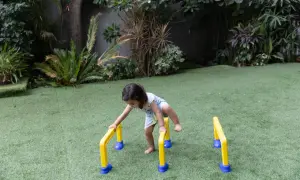 A toddler practices gross motor outdoor activities, balancing on yellow and blue hurdles in a grassy yard.
