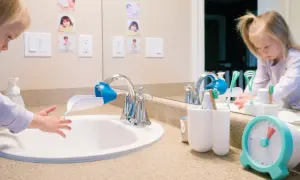 A child washes hands at a sink with a faucet extender, in a Montessori bathroom. Toothbrushes and a timer are nearby.