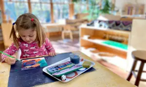 A young girl with pigtails paints at a table, exploring her creativity with watercolors. This scene captures the essence of Montessori nature-inspired learning.
