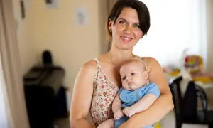 A smiling mother holds her 2-month-old baby, celebrating precious milestones. The baby wears a blue onesie with a yellow and blue heart.