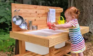 A toddler engages in Montessori movement activities, exploring a wooden mud kitchen with pots and a watering can.