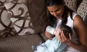 A mother gently feeds her baby from a bottle, holding him in a comfortable, paced feeding position on her lap.