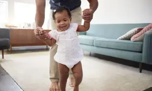 A father helps his baby practice gross motor activities for infants 12-18 months, taking first steps with a smile.