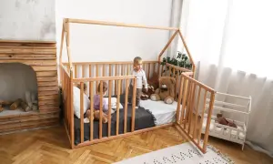 Two children play in a wooden playpen floor bed. One reads a book, the other stands, with a teddy bear nearby.