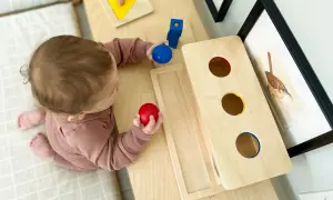 A baby explores Montessori activities for 10 month old baby, holding red and blue balls near a wooden drop box and shape puzzle.