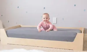 A smiling baby in pink pajamas explores a Montessori bed with a grey mattress. The wooden bed frame is low to the ground, perfect for little adventurers.