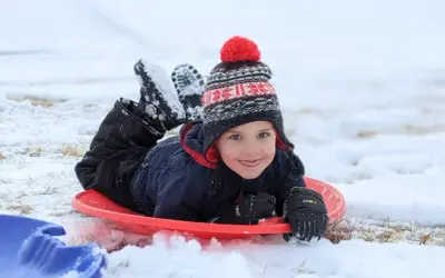 A happy child in a winter hat and snowsuit lies on a red sled, ready for fun. Perfect for things to do with baby in winter, like sledding!