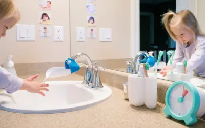 A child washes hands at a sink with a faucet extender, in a Montessori bathroom. Toothbrushes and a timer are nearby.
