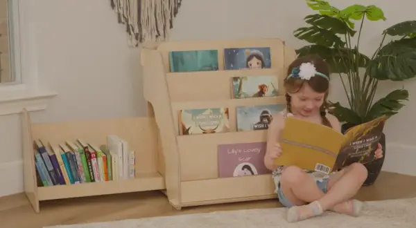 A young girl reads a book next to a DIY Montessori bookshelf filled with children's books.