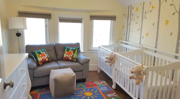 A charming nursery with two white cribs, perfect for nursery sets for twins. A grey sofa with fox-themed pillows and a colorful rug complete the cozy space.