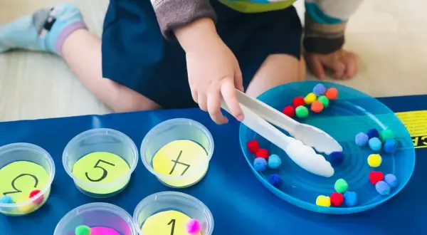 Toddler sorting colorful pom-poms into numbered cups. A fun activity for what to do with toddlers at home, practicing fine motor skills and number recognition.
