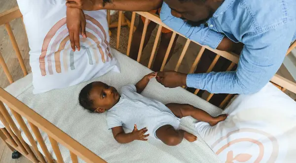 Parents hold their baby's hand in a crib with a breathable mattress, a safe sleep environment.