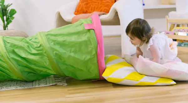 A toddler explores a green play tunnel, engaging in fun climbing activities. Colorful pillows add to the playful scene.