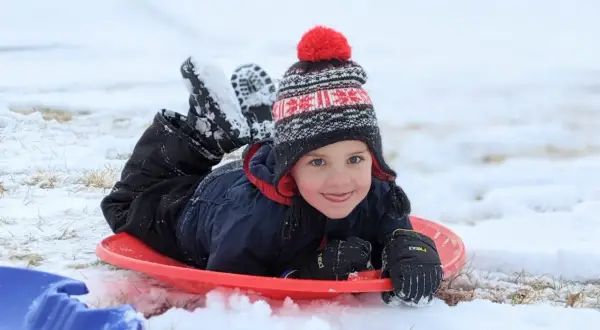 A happy child in a winter hat and snowsuit lies on a red sled, ready for fun. Perfect for things to do with baby in winter, like sledding!