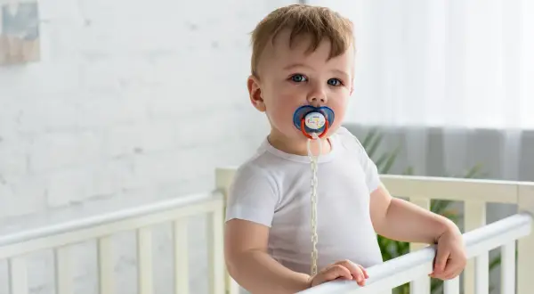 A baby in a crib with a pacifier. Some wonder if a pacifier can cause speech delay, but this little one seems content.