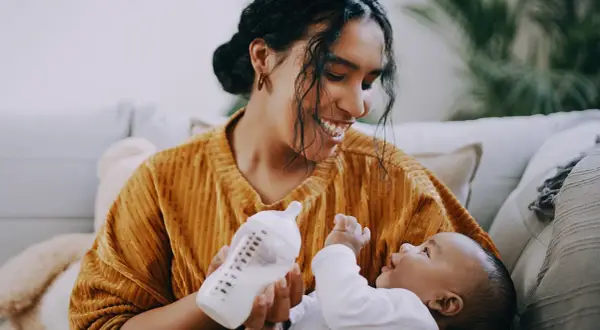 A mother smiles while paced feeding her newborn with a bottle. The baby looks up at her, reaching out a hand.