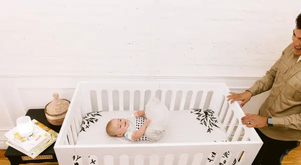 A happy baby rolls around in a white baby crib twin. A man smiles as he looks into the crib.
