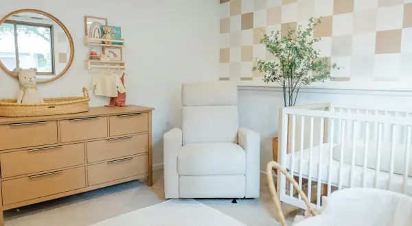 Cozy and modern nursery with a wooden dresser, plush recliner, and crib. This room offers sweet, cute baby room ideas with its neutral palette and playful checkered wallpaper.