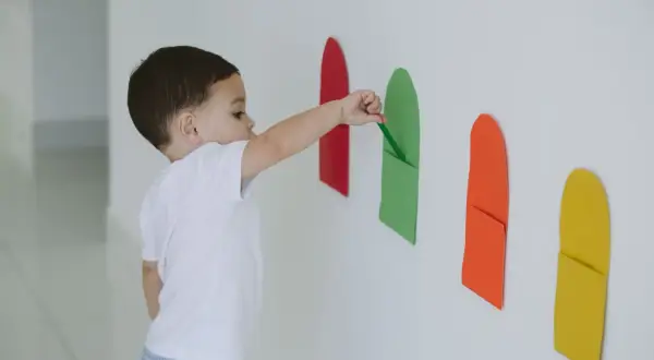 A toddler explores bright colors for 1 year old, placing a green paper strip into a matching pocket on a white wall.