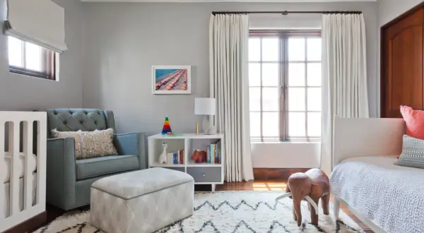 A cozy nursery features a tufted blue couch, a crib, and a white bookshelf filled with toys and books.