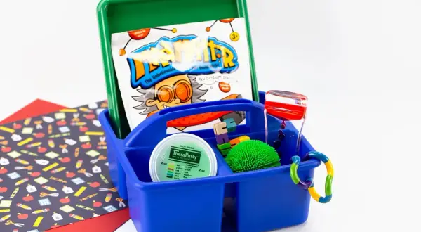 A blue caddy holds items for a sensory box: TheraPutty, a liquid timer, a spiky ball, and building blocks.