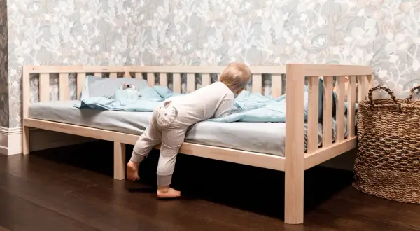 A toddler climbs into a Montessori bed, showcasing its low profile and ideal dimensions for independent exploration.