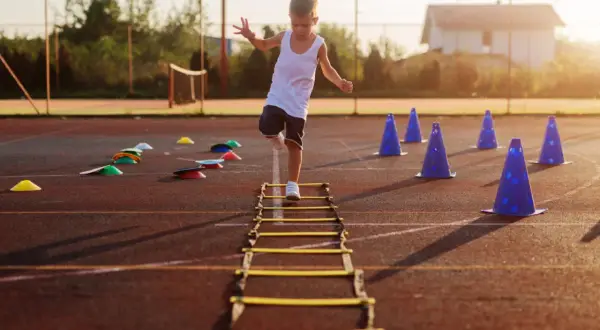 A young boy practices gross motor activities for 5 year olds, stepping through an agility ladder on a sports field with cones.