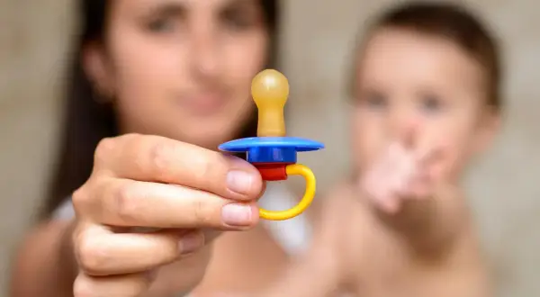 A mother holds a colorful pacifier, perhaps contemplating extended pacifier use for her baby, who is blurred in the background.