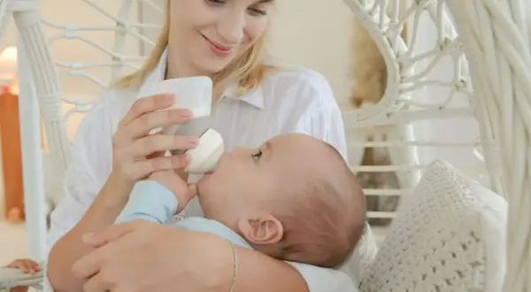 A mother smiles lovingly while holding her baby for upright bottle feeding. The baby is nestled in her arms, taking milk from the bottle.