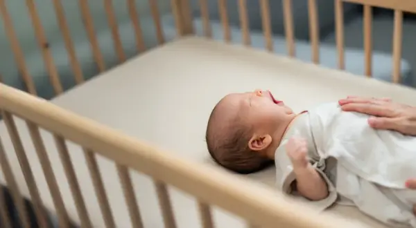 A baby yawns in a wooden crib. This image reassures parents that mini cribs are safe for their little ones.