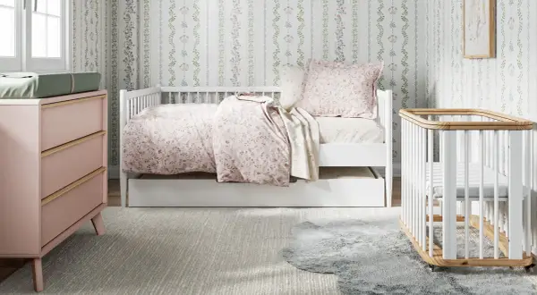 A charming small nursery features a white daybed with floral bedding, a pink dresser, and a white crib.