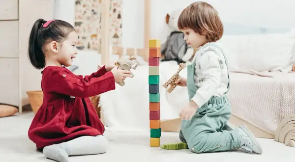 Two toddlers engage in fun games to play with one year old, stacking colorful blocks and playing with wooden animals.