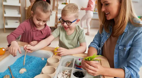 Children explore blue sand and shells in a sensory table, guided by a smiling teacher.