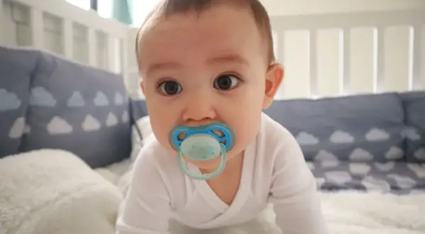 A baby with a blue orthodontic pacifier in a crib. Are orthodontic pacifiers better for developing smiles?
