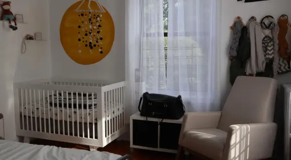 Cozy nursery nook ideas: a white crib, a comfy armchair, and a wall decorated with a sun mural and a New York pennant.