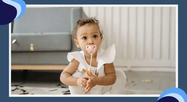 A baby girl with a pink pacifier in her mouth sits on the floor, her eyes wide with curiosity. The pacifier terth is a comforting presence as she explores her surroundings.